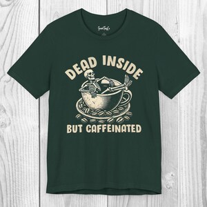 Può includere: T-shirt verde scuro con una grafica raffigurante uno scheletro che si rilassa in una tazza di caffè circondato da chicchi di caffè. Il testo recita "DEAD INSIDE BUT CAFFEINATED" in un font in stile vintage.