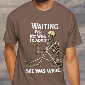 May include: Brown t-shirt with a humorous graphic. The design features a skeleton sitting and holding an hourglass, with the text "WAITING FOR MY WIFE TO ADMIT SHE WAS WRONG". A butterfly and flowers are also included in the design.