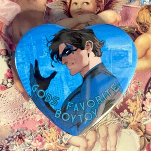 Puede incluir: Un botón en forma de corazón con fondo azul. Presenta una ilustración de cómic de un personaje con un traje azul y negro. El texto "GODS FAVORITE BOYTOY" está impreso en el botón.