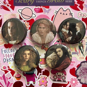 May include: Five circular pin-back buttons featuring photographic scenes from the film Interview with the Vampire. The buttons display portraits of characters with long hair, period clothing, and dramatic expressions, arranged on a pink background.