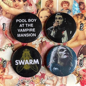 May include: Four black and white button pins with different designs. The first pin says "Pool Boy at the Vampire Mansion". The second pin features a black and white photo of a person with a microphone. The third pin has a black and white illustration of a fly with the word "Swarm" printed below it. The fourth pin features a black and white photo of a person with long hair.