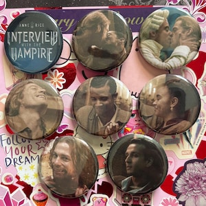 May include: A collection of eight circular pin-back buttons featuring imagery from Interview with the Vampire. One button displays the movie title text, while the others show various character portraits and scenes from the film in a sepia-toned style.