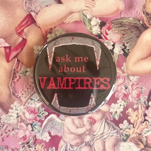 Peut inclure: Un bouton rond noir avec le texte rouge "ask me about VAMPIRES". Le bouton présente un dessin stylisé de crocs de vampire. Le fond est un motif floral rose.