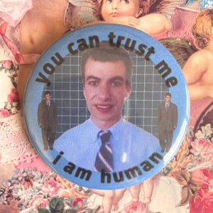May include: A blue and white button with the text "You can trust me I am human" in black lettering. The button features a photo of a man in a blue shirt and a striped tie.