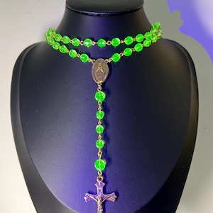 Könnte beinhalten: Eine neongrüne Rosenkranzkette mit einem goldfarbenen Kruzifix und einem Anhänger mit einer religiösen Figur. Die Perlen sind leuchtend grün, präsentiert auf einer schwarzen Schaufensterpuppe.