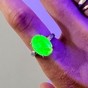 Könnte beinhalten: Ein Silberring mit einem großen, ovalen, leuchtend neongrünen Edelstein, der unter ultraviolettem Licht intensiv strahlt. Die Metallschiene ist auf beiden Seiten der zentralen ovalen Fassung mit kleinen, klaren, runden Schmucksteinen besetzt.