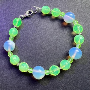 Può includere: Un braccialetto di perline con una miscela di perline sfaccettate verde neon e blu opalescente. Il braccialetto è infilato su una catena d'argento e ha una chiusura d'argento. Le perline sono rotonde e lisce, creando un accessorio vivace.