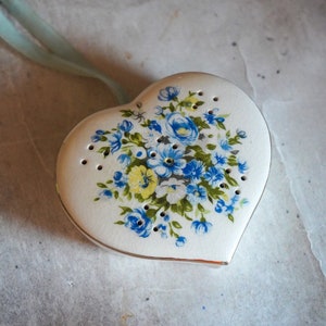 Op de afbeelding: Een wit hartvormig keramiek ornament met een blauw en geel bloemenontwerp. Het ornament heeft kleine gaatjes over het hele oppervlak.