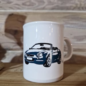 May include: White ceramic mug with a black and blue illustration of a convertible car. The car has a black interior and a black top.