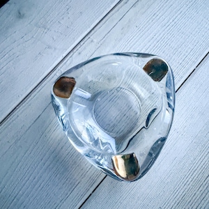 Puede incluir: Cenicero de vidrio transparente con forma triangular y tres apoyos dorados. El cenicero tiene una hendidura circular en el centro y está sobre una superficie de madera clara.
