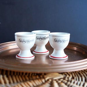 以下が含まれることがあります： 赤色のベースと黒い筆記体で「Guten Appetit」の文字が入った白いエッグカップが3つ。エッグカップは銅色のトレイの上に置かれ、濃い青色の背景をしています。