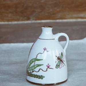 Könnte beinhalten: Ein kleiner weißer Keramik Krug mit braunem Rand und Henkel. Der Krug ist mit einer grünen Libelle, rosa Blumen und grünen Blättern verziert.