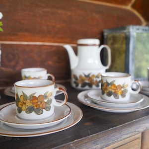 Peut inclure: Un ensemble de tasses et soucoupes à thé blanches avec un motif floral dans des tons de brun, jaune et orange. Les tasses et les soucoupes sont disposées sur une table en bois avec une théière blanche en arrière-plan.