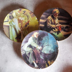 Może przedstawiać: Trzy dekoracyjne ceramiczne talerze z malowanymi obrazami kobiet w odzieży vintage. Talerze są okrągłe i mają złotą obwódkę. Pierwszy talerz przedstawia kobietę w białej sukience z czerwoną chustką, drugi talerz przedstawia kobietę w brązowej kurtce i zielonej spódnicy, która dzierga, a trzeci talerz przedstawia kobietę w białej sukience, która miesza garnek nad ogniem.