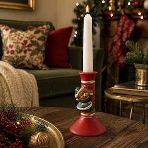 Puede incluir: Un candelabro rojo con un oso de peluche esculpido que lleva un gorro y un traje verdes. El oso está colocado en la columna central del soporte, que tiene detalles en color dorado. Una vela blanca cónica está encendida y colocada dentro.