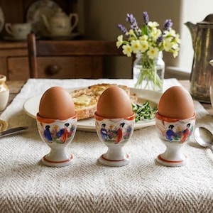 May include: Three brown eggs in decorative egg cups with a painted design, set on a white cloth. A breakfast scene with toast, butter, honey, and a silver pitcher. A vintage aesthetic.