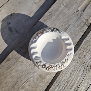 Puede incluir: Cenicero de cerámica blanca con un diseño floral alrededor del borde. El cenicero tiene cuatro reposacigarrillos y está sobre una superficie de madera envejecida. El diseño floral incluye detalles dorados y plateados.