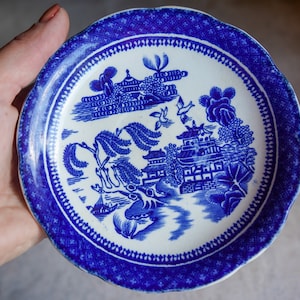 May include: A small, white ceramic saucer with a blue and white willow pattern. The saucer has a scalloped edge and a blue floral border.