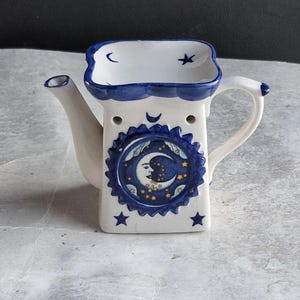 Könnte beinhalten: Ein weißer, keramik-teekannenförmiger Ölwärmer mit blauem und weißem Himmelsdesign. Der Wärmer hat einen Mond und Sterne, einen Griff und einen Ausguss. Die Oberseite hat eine kleine Schale mit blauem Rand und einem Mond und Stern.