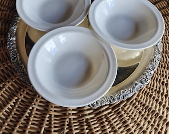 May include: Three white ceramic bowls with a simple design are arranged on a silver-colored tray with an ornate edge. The tray rests on a woven wicker surface, creating a contrast of textures and colors.
