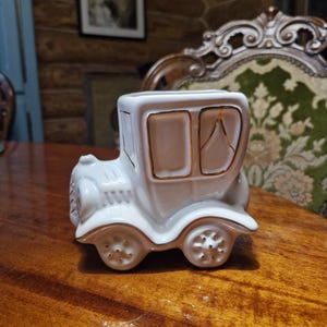Pode incluir: Uma peça decorativa em cerâmica branca em forma de carruagem com detalhes dourados. A carruagem em estilo vintage tem duas janelas e quatro rodas. A peça está sobre uma superfície de madeira.