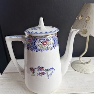 Puede incluir: Cafetera de cerámica blanca con tapa, con diseños florales y geométricos en rojo, azul y amarillo. La cafetera tiene asa y boquilla, colocada sobre una superficie de madera blanca.