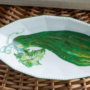 May include: White, octagonal serving dish with a green rim and a painted cucumber design. The cucumber is green with yellow highlights and is accompanied by green leaves and vines. The dish is on a woven wicker surface.