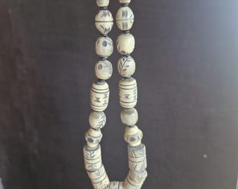May include: Cream-colored beaded necklace with etched black designs. The necklace features a variety of bead shapes, including round and cylindrical, strung together with a silver clasp. The beads have a natural, organic appearance.