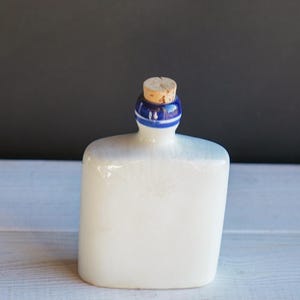 May include: A white ceramic flask with a cork stopper. The neck of the flask is adorned with a blue and white striped design. The flask has a rectangular shape with rounded edges, and it sits on a white wooden surface.