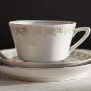 May include: A white teacup and saucer set with a delicate silver floral pattern around the cup's rim. The cup sits on a matching saucer and a larger plate, creating a layered effect. The set is on a white surface.