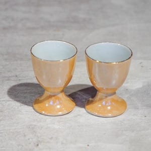 Pode incluir: Duas porta-ovos iridescentes com acabamento perolado. Os porta-ovos têm uma base alargada e uma borda dourada. O interior das chávenas é azul claro. Os porta-ovos estão sobre uma superfície cinzenta.