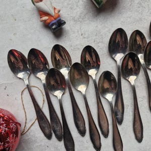 Könnte beinhalten: Eine Sammlung von Vintage-Silberlöffeln mit länglichen Griffen, angeordnet auf einer hellen Oberfläche. Eine rote Weihnachtskugel und eine kleine Zwergfigur sind ebenfalls vorhanden. Die Löffel haben eine reflektierende Oberfläche.