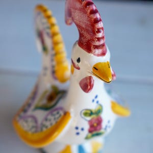 Puede incluir: Figura de gallo de cerámica con cuerpo blanco, cresta roja y pico amarillo. Las alas y la cola presentan patrones florales coloridos con detalles amarillos y azules. Un objeto decorativo.