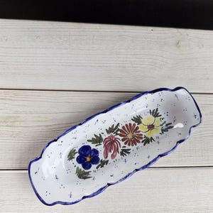 Könnte beinhalten: Eine weiße Keramik-Servierschale mit gewelltem Rand und blauem Rand. Die Schale ist mit bunten, handgemalten Blumen in Blau-, Rot- und Gelbtönen verziert. Die Schale ist etwa 30 cm lang.