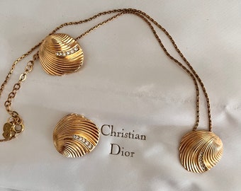 May include: A gold necklace with a shell pendant and matching earrings. The pendant and earrings are adorned with small clear stones. The necklace is on a white background with the text "Christian Dior" visible.