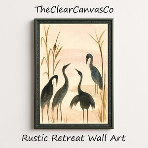 May include: A framed watercolor painting featuring four dark gray cranes in a marsh setting. The artwork has a neutral color palette with beige, brown, and pink tones. The text "TheClearCanvasCo" and "Rustic Retreat Wall Art" are visible.