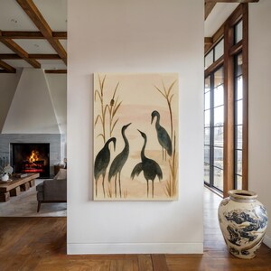 May include: A canvas art print featuring a watercolor painting of five dark gray cranes in a marsh setting. The background has a soft pink and beige color palette with tall reeds. The artwork is displayed on a white wall.