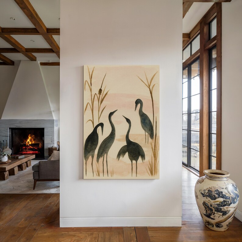 May include: A canvas art print featuring a watercolor painting of five dark gray cranes in a marsh setting. The background has a soft pink and beige color palette with tall reeds. The artwork is displayed on a white wall.