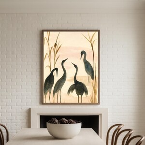 May include: Watercolor painting of four dark blue herons in a natural setting, framed in a brown wooden frame. The background features a soft pink and beige sky with tall, thin reeds. The artwork is displayed above a white fireplace.