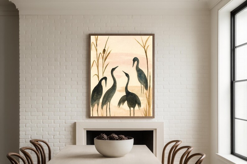 May include: Watercolor painting of four dark blue herons in a natural setting, framed in a brown wooden frame. The background features a soft pink and beige sky with tall, thin reeds. The artwork is displayed above a white fireplace.