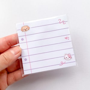 Op de afbeelding: Een vierkant notitieblok met wit papier met horizontale lichtpaarse lijnen en een verticale roze kantlijn. Het papier bevat kleine illustraties van een bruin puppygezicht, een roze hartje en een wit en roze kattengezicht, verpakt in plastic.
