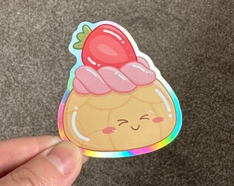 Op de afbeelding: Een holografische sticker van een cartoon gebakje met een roze swirl en een rode aardbei erop. Het gebakje heeft een glimlachend gezicht.