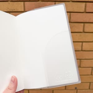 Op de afbeelding: Open wit notitieboek met een doorzichtige plastic zak. De zak heeft een kleine illustratie van een hond en een kat, met de tekst "CUTE STICKERS ONLY" eronder. Het notitieboek is open en toont lege pagina's.