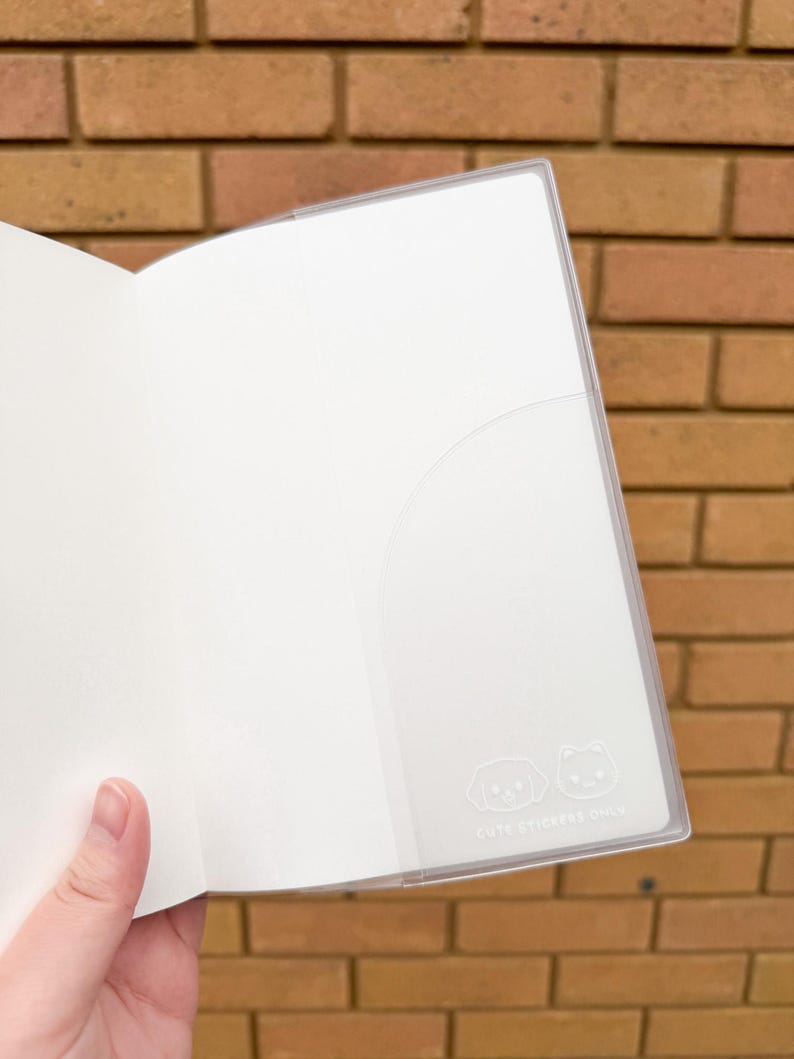 Op de afbeelding: Open wit notitieboek met een doorzichtige plastic zak. De zak heeft een kleine illustratie van een hond en een kat, met de tekst "CUTE STICKERS ONLY" eronder. Het notitieboek is open en toont lege pagina's.