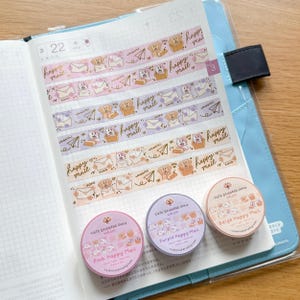 May include: Three rolls of washi tape with cute illustrations of dogs and envelopes. The tape is labelled "Pink Happy Mail", "Purple Happy Mail", and "Beige Happy Mail".