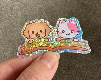Op de afbeelding: Een glinsterende sticker met een cartoonhond en -kat die samen een boek lezen. Het boek is open en de dieren kijken over de bovenkant van de pagina's. De sticker heeft een holografische afwerking.