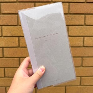 以下が含まれることがあります： 「the YEARLY NOTEBOOK」の文字が入ったグレーのノート。透明なプラスチックカバー付き。レンガの壁を背景にノートが手に持たれています。「ONE YEAR EDITION」の文字が下部に印刷されています。
