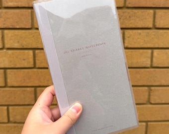 Puede incluir: Un cuaderno gris con el texto "the YEARLY NOTEBOOK" en una cubierta de plástico transparente. El cuaderno se sostiene frente a una pared de ladrillos. Las palabras "ONE YEAR EDITION" están impresas en la parte inferior.