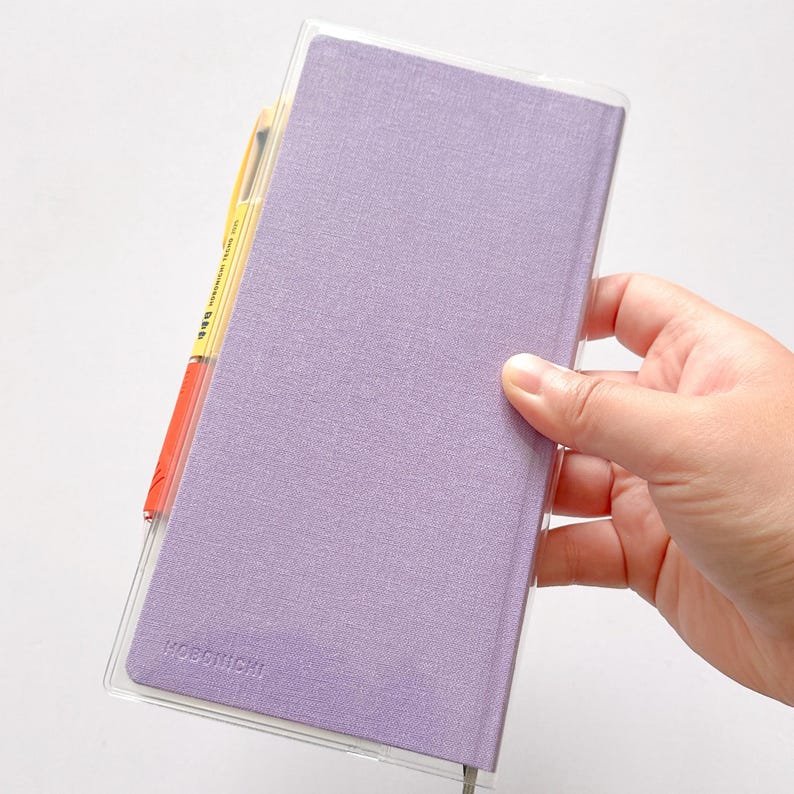 Op de afbeelding: Een lavendelkleurige Hobonichi-planner met een transparante plastic hoes. De planner wordt in een hand gehouden.
