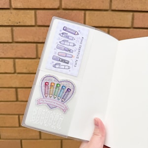Op de afbeelding: Een doorzichtig plastic notitieboek met stickerbladen. E&eacute;n vel bevat illustraties van pennen, de andere heeft een hartvormig ontwerp met regenboogkleurige penillustraties en de tekst "My Favourite Rainbow". De tekst "Cute Stickers Only" is ook zichtbaar.
